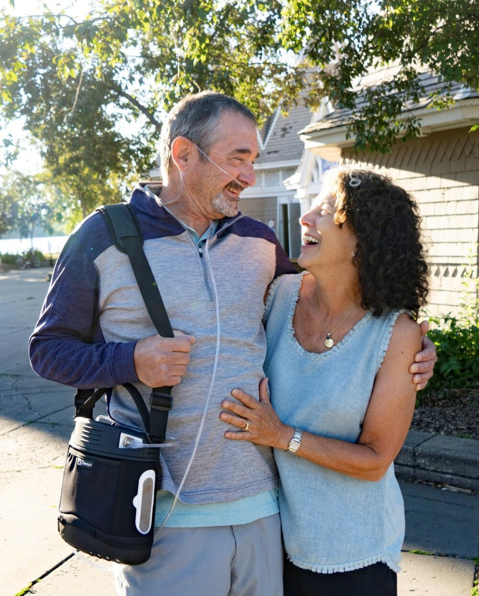 A couple is walking and smiling. The man is carrying a Oxlife LIBERTY™ Portable Oxygen Concentrator in a carry bag over his shoulder
