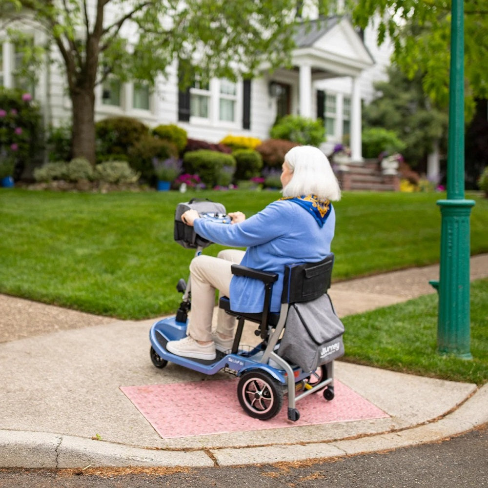 An older woman riding a Blue Journey So Lite Foldable Power Scooter onto a curbside pavement.